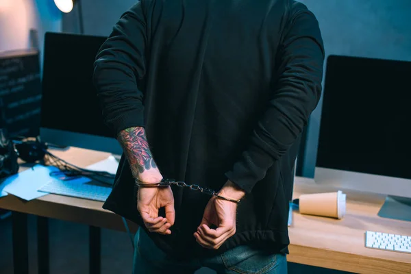 Cropped shot of handcuffed hacker in hoodie in front of his work desk — Stock Photo