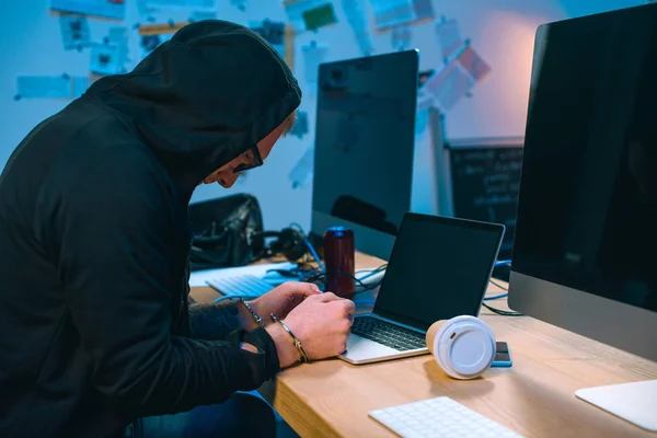 Hacker esposado con capucha delante de la computadora portátil en el lugar de trabajo - foto de stock