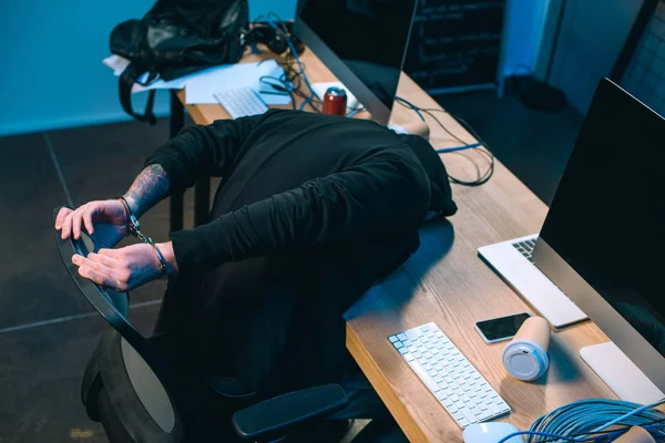 Handcuffed hacker in hoodie leaning on his work desk — Stock Photo