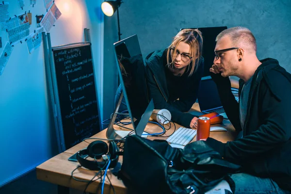 Casal pensativo de hackers olhando para a tela do computador juntos no quarto escuro — Fotografia de Stock