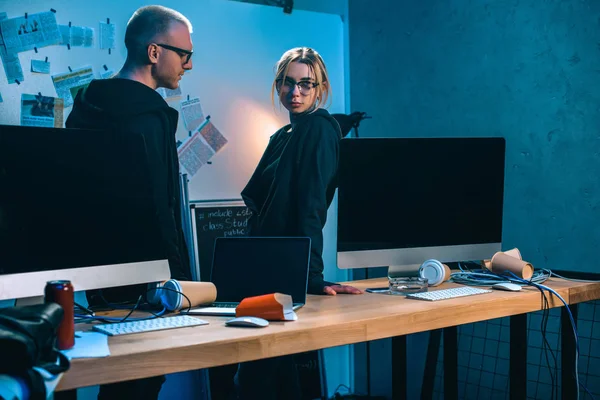 Couple of hackers standing near desk with computers in dark room — Stock Photo