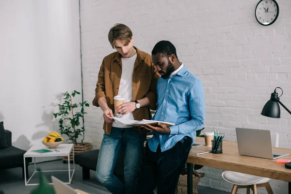 Hombre de negocios con taza de papel de café viendo en libro de texto y hablando con compañero de trabajo afroamericano - foto de stock