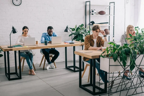 Colegas de negocios multiculturales sentados a la mesa y trabajando en computadoras portátiles en la oficina moderna - foto de stock