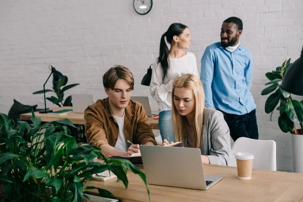 Colleghi di lavoro che scrivono in un libro di testo e colleghi in piedi dietro in ufficio moderno — Foto stock