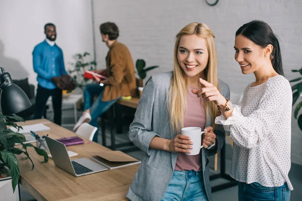 Sonriente mujer de negocios señalando con el dedo a su colega y dos compañeros de trabajo de pie detrás en la oficina moderna - foto de stock