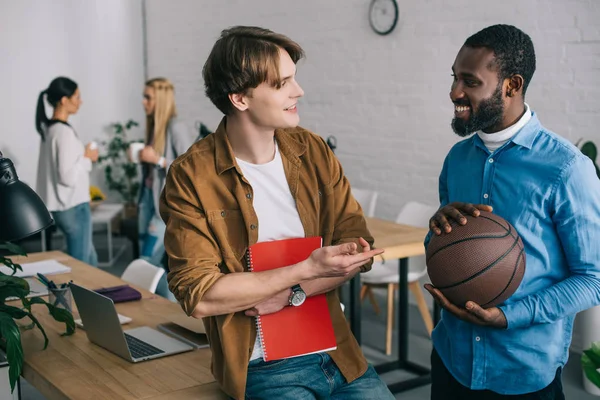 Sonriente hombre de negocios afroamericano sosteniendo la pelota y hablando con su pareja con el libro de texto y dos mujeres de negocios de pie detrás con café - foto de stock