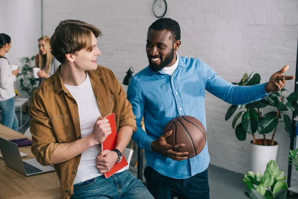 Hombre de negocios afroamericano con bola señalando con el dedo a colega y mujeres de negocios de pie detrás con tazas de café - foto de stock