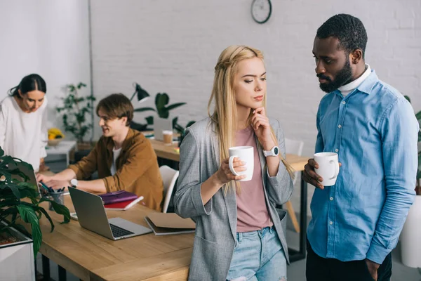 Compañeros de negocios multiculturales con tazas de café teniendo discusión y dos compañeros de trabajo que trabajan en el ordenador portátil detrás - foto de stock