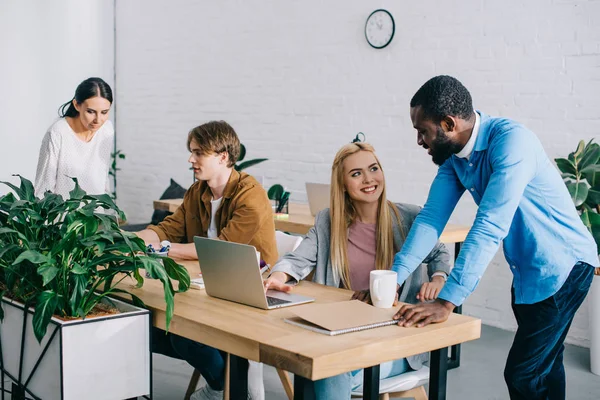 Homme d'affaires afro-américain parlant à une collègue et deux collègues travaillant à table avec des ordinateurs portables — Stock Photo