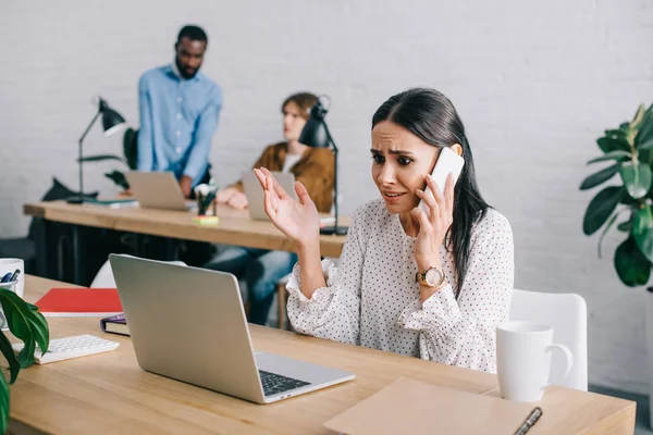 Molesto mujer de negocios hablando en el teléfono inteligente y colegas que trabajan detrás en la oficina moderna - foto de stock