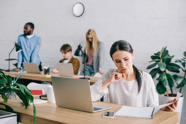 Jovem empresária assistindo no livro didático e colegas de trabalho que trabalham para trás no escritório moderno — Fotografia de Stock