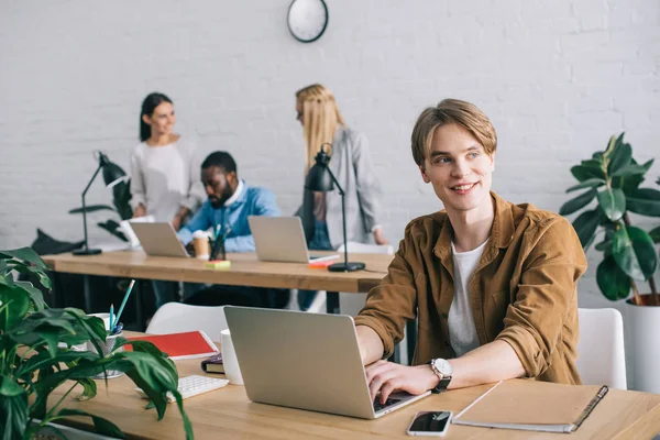 Smiling businessman with laptop and colleagues working behind in modern office — Stock Photo