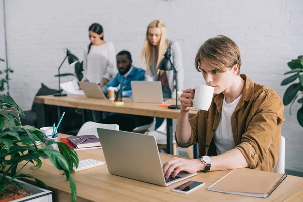 Homem de negócios com caneca de café usando laptop e colegas que trabalham para trás no escritório moderno — Fotografia de Stock