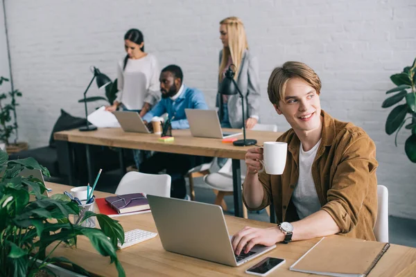 Homem de negócios com caneca de café usando laptop e colegas que trabalham para trás no escritório moderno — Fotografia de Stock