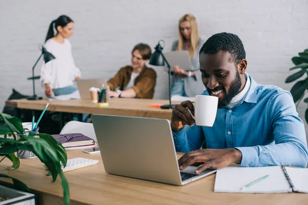 Sonriente hombre de negocios afroamericano utilizando el ordenador portátil y beber café de taza y compañeros de trabajo que trabajan detrás en la oficina moderna - foto de stock