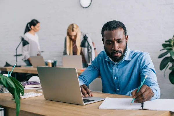 Joven empresario afroamericano escribiendo en libro de texto y usando portátil en la mesa y dos mujeres de negocios que trabajan detrás — Stock Photo