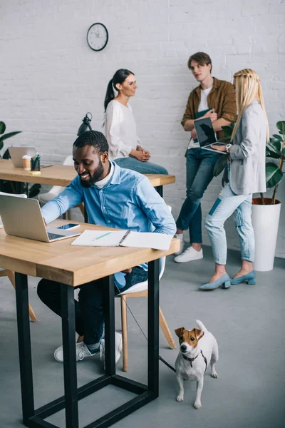 Lächelnder afrikanisch-amerikanischer Geschäftsmann, der am Laptop arbeitet und Hund an der Leine hält und Mitarbeiter, die sich hinter ihm treffen — Stockfoto