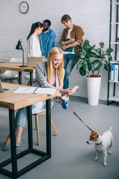 Mulher de negócios sorridente que tira foto do cão em coleira e colegas de trabalho que se encontram atrás no escritório moderno — Fotografia de Stock