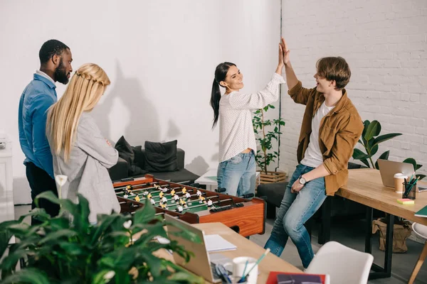 Smiling business coworkers celebrating victory in table football and taking high five to each other in front of losing colleagues — Stock Photo