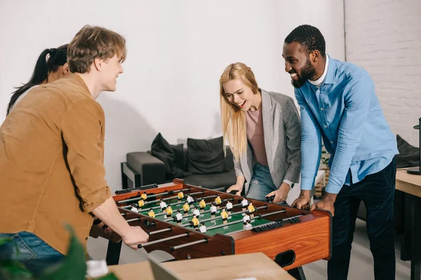 Sonrientes colegas de negocios multiétnicos jugando futbolín en la oficina moderna - foto de stock