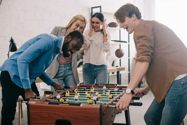 Vista lateral de empresarios multiculturales felices jugando al futbolín frente a colegas mujeres - foto de stock