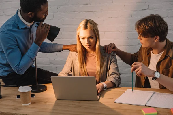 Dos hombres de negocios multiculturales que amenazan y brillan lámpara en la cara de la mujer de negocios utilizando el ordenador portátil - foto de stock