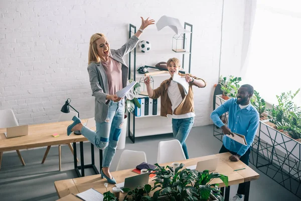 Vue d'angle élevé des collègues d'affaires multiethniques heureux jetant des papiers et s'amusant dans le bureau moderne — Photo de stock