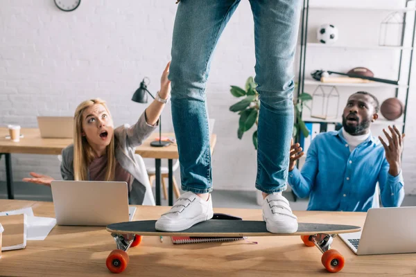 Imagen recortada de hombre de negocios montando en longboard en la mesa y compañeros conmocionados en la oficina moderna - foto de stock