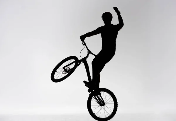 Silhouette eines Trial-Bikers, der auf dem Hinterrad steht und die Hand auf weiß hebt — Stockfoto