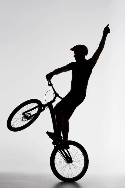 Silueta del ciclista de prueba de pie en la rueda trasera y levantando la mano en blanco — Stock Photo