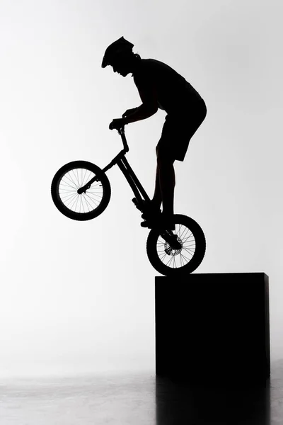 Silhueta de julgamento motociclista realizando acrobacia ao equilibrar no cubo no branco — Fotografia de Stock