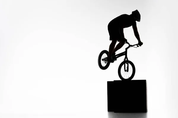 Silhouette du cycliste d'essai effectuant cascade tout en équilibrant sur cube sur blanc — Photo de stock