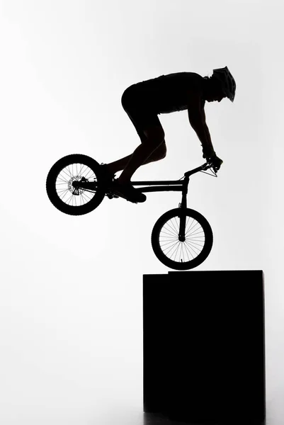 Silhouette of trial cyclist performing nollie while balancing on cube on white — Stock Photo