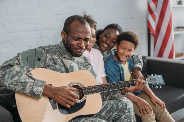 African american woman and children listening to father in camouflage clothes playing guitar — Stock Photo