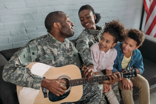 Mujer soldado afroamericana y niños escuchando a su padre en ropa de camuflaje tocando la guitarra - foto de stock