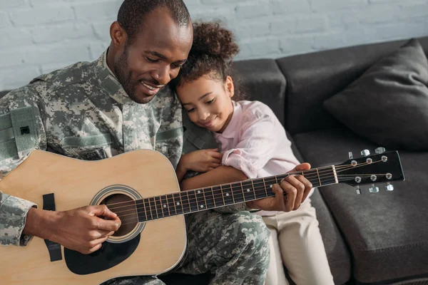 Sonriente soldado tocando la guitarra y abrazando a su hija - foto de stock