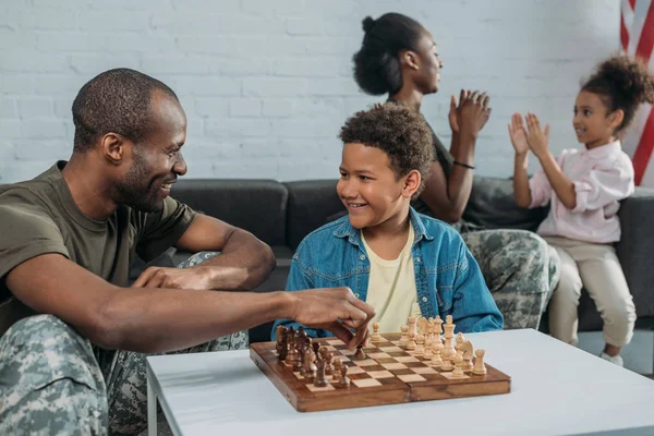 Soldat souriant jouant aux échecs avec son fils tandis que la mère et la fille jouent ensemble — Photo de stock