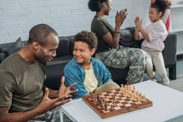 Homme en tenue de camouflage enseignant à son fils à jouer aux échecs pendant que mère et fille jouent ensemble — Photo de stock