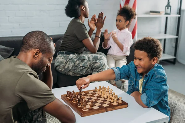 Père en uniforme de l'armée jouant aux échecs avec son fils pendant que la mère et la fille jouent ensemble — Photo de stock