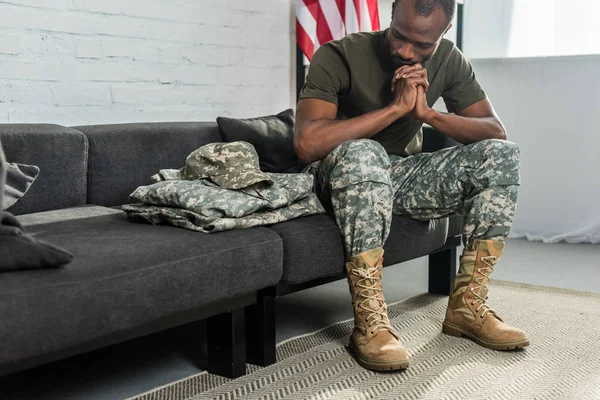 Задумчивый солдат сидит на диване и смотрит на свою камуфляжную одежду — стоковое фото