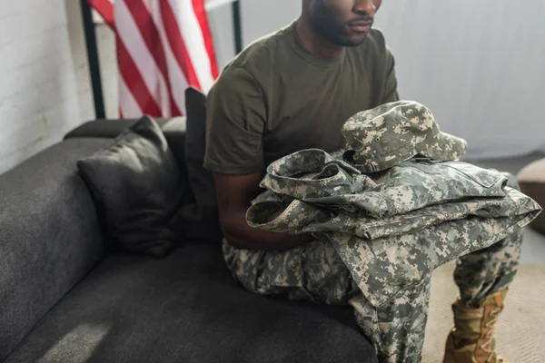 Guapo soldado sosteniendo su ropa de camuflaje mientras está sentado en el sofá - foto de stock