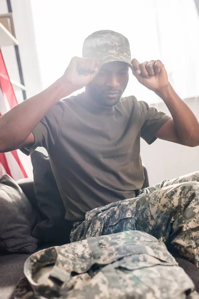 Guapo soldado poniéndose la gorra mientras está sentado en el sofá - foto de stock