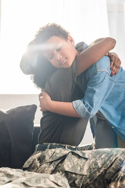 Female army soldier in camouflage clothes and her son embracing on sofa — Stock Photo