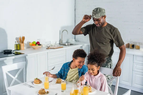 Отец солдат армии смотрит на счастливых детей, наслаждающихся едой на кухне — стоковое фото