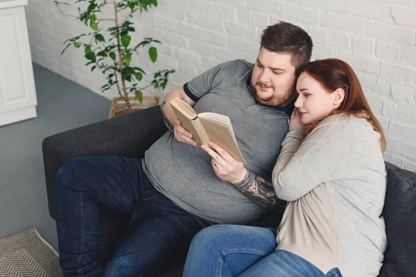 Novio y novia leyendo libro juntos en el sofá en la sala de estar - foto de stock