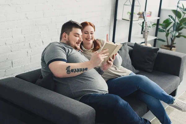 Улыбающиеся толстый парень и девушка читают книгу на диване дома — стоковое фото