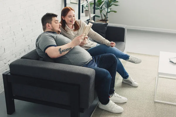 Novio y novia leyendo libro juntos en el sofá en casa - foto de stock