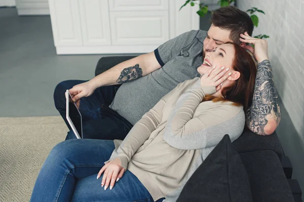 Смеющиеся парень и девушка лежат на диване и смотрят что-то на планшете дома — стоковое фото