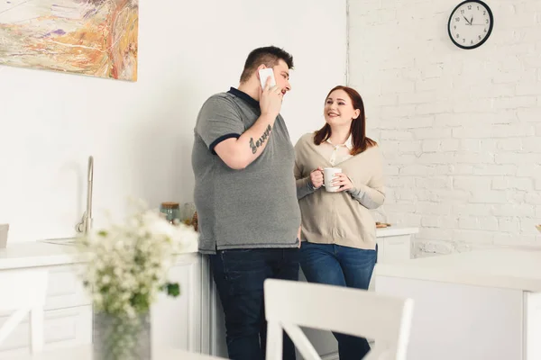 Boyfriend talking by smartphone and happy girlfriend holding cup of coffee in kitchen — Stock Photo