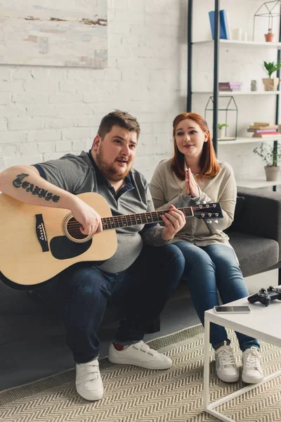 Девушка спрашивает парня играть песню с акустической гитарой дома — стоковое фото